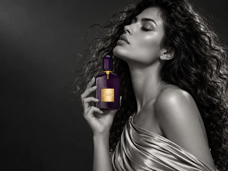 Editorial illustration: a black and white portrait of a woman with long curls holding a small purple fragrance bottle near her face