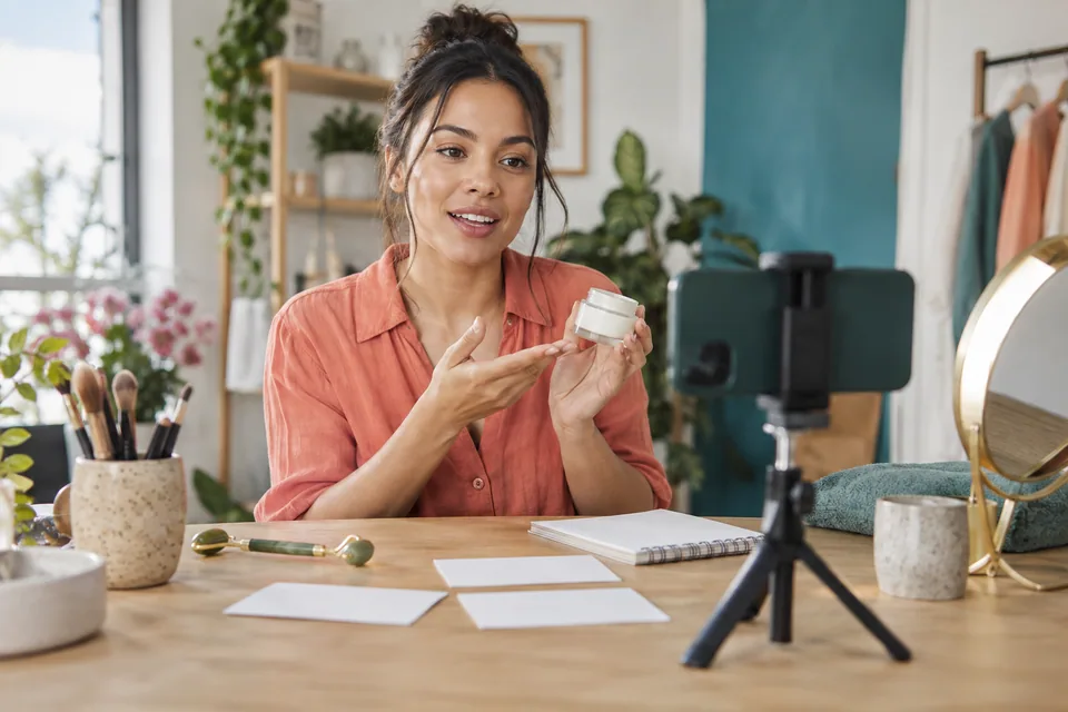 Editorial illustration: A fictional adult beauty creator in a bright home studio recording a simple skincare product explanation using a smartphone on a small tripod, an unbranded cream jar, reflector and handwritten-looking blank storyboard cards