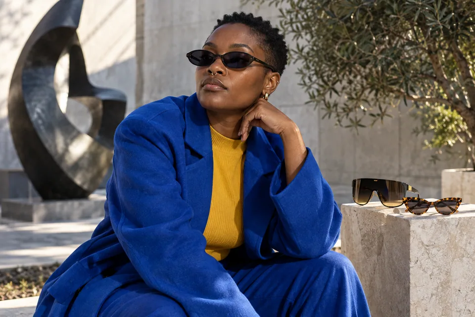 Editorial illustration: An expressive fictional Black nonbinary adult wearing slim oval acetate sunglasses at a contemporary sculpture courtyard, oversized shield and updated cat-eye frames subtly displayed on a nearby small pedestal